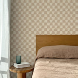 Bed with tan linens and wooden headboard, wooden side table  with white book and green glass, white floor length curtain against a watercolor checkerboard wallpaper in warm beige and clay brown.