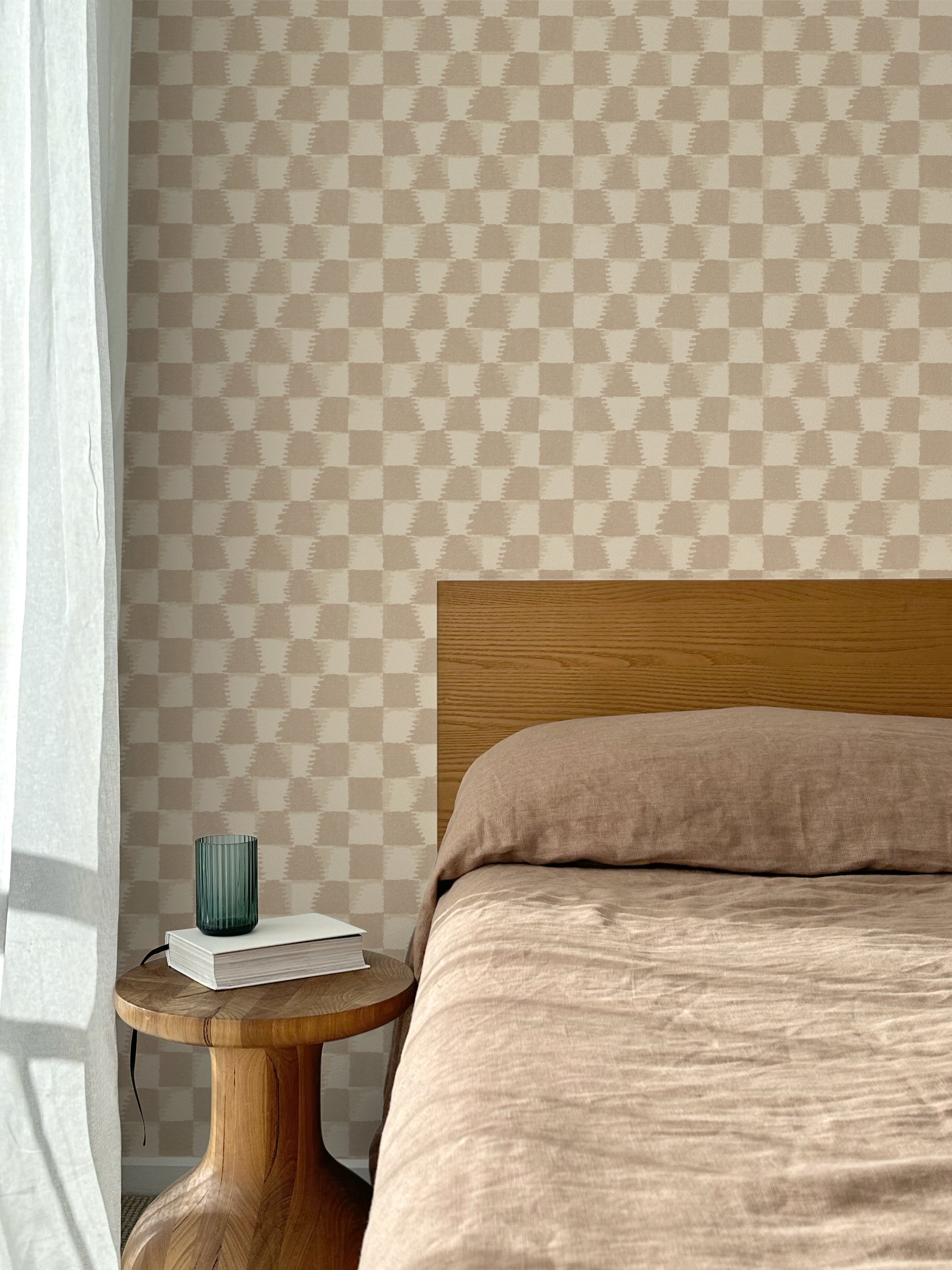 Bed with tan linens and wooden headboard, wooden side table  with white book and green glass, white floor length curtain against a watercolor checkerboard wallpaper in warm beige and clay brown.