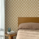 Bed with tan linens and wooden headboard, wooden side table  with white book and green glass, white floor length curtain against a watercolor checkerboard wallpaper in tan and beige.