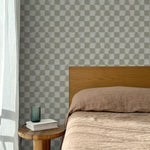 Bed with tan linens and wooden headboard, wooden side table  with white book and green glass, white floor length curtain against a watercolor checkerboard wallpaper in taupe-grey. 
