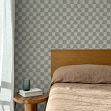 Bed with tan linens and wooden headboard, wooden side table  with white book and green glass, white floor length curtain against a watercolor checkerboard wallpaper in taupe-grey. 