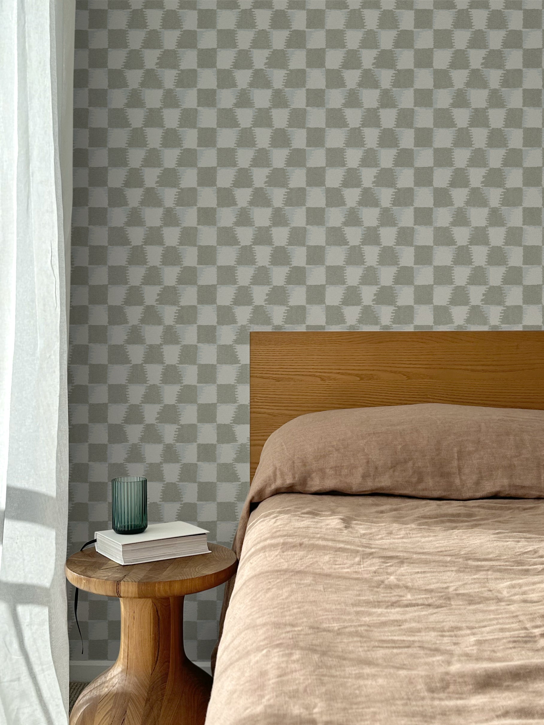 Bed with tan linens and wooden headboard, wooden side table  with white book and green glass, white floor length curtain against a watercolor checkerboard wallpaper in taupe-grey. 
