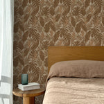 Bed with tan linens and wooden headboard, wooden side table  with white book and green glass, white floor length curtain against a brown tropical wallpaper. 