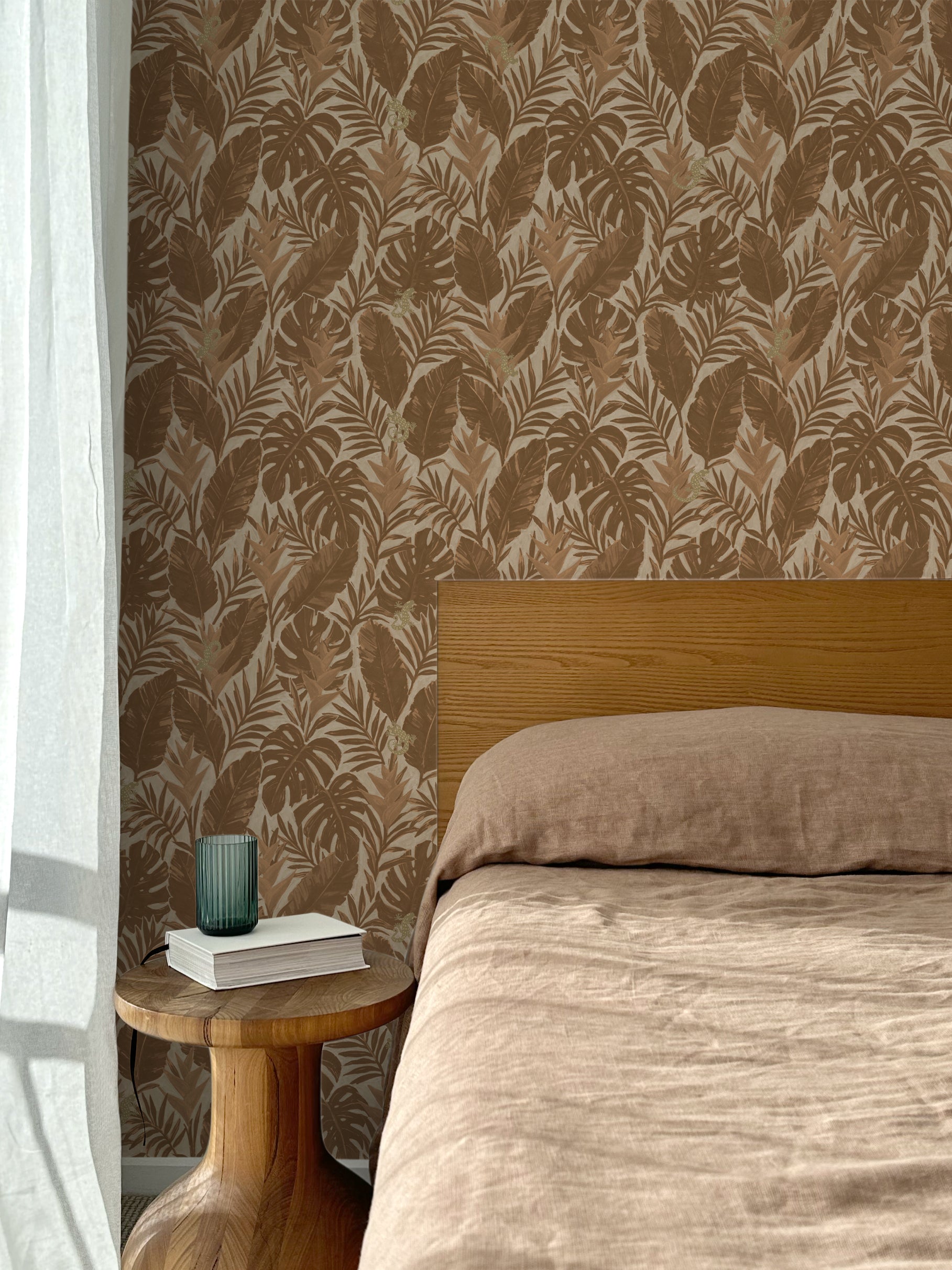 Bed with tan linens and wooden headboard, wooden side table  with white book and green glass, white floor length curtain against a brown tropical wallpaper. 