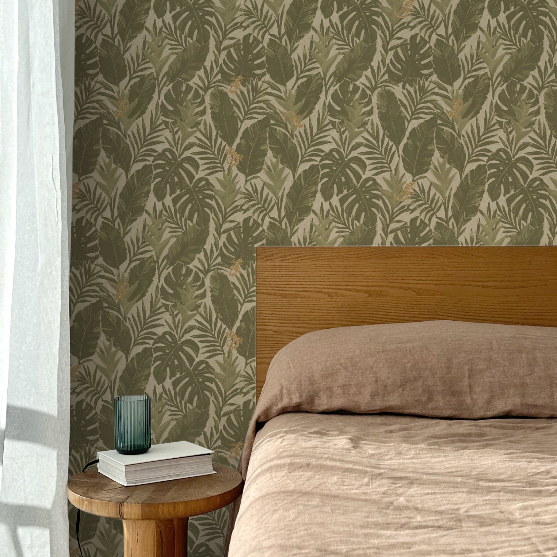 Bed with tan linens and wooden headboard, wooden side table  with white book and green glass, white floor length curtain against a olive green and tan tropical wallpaper. 
