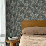 Bed with tan linens and wooden headboard, wooden side table  with white book and green glass, white floor length curtain against a steel blue and tan tropical wallpaper. 