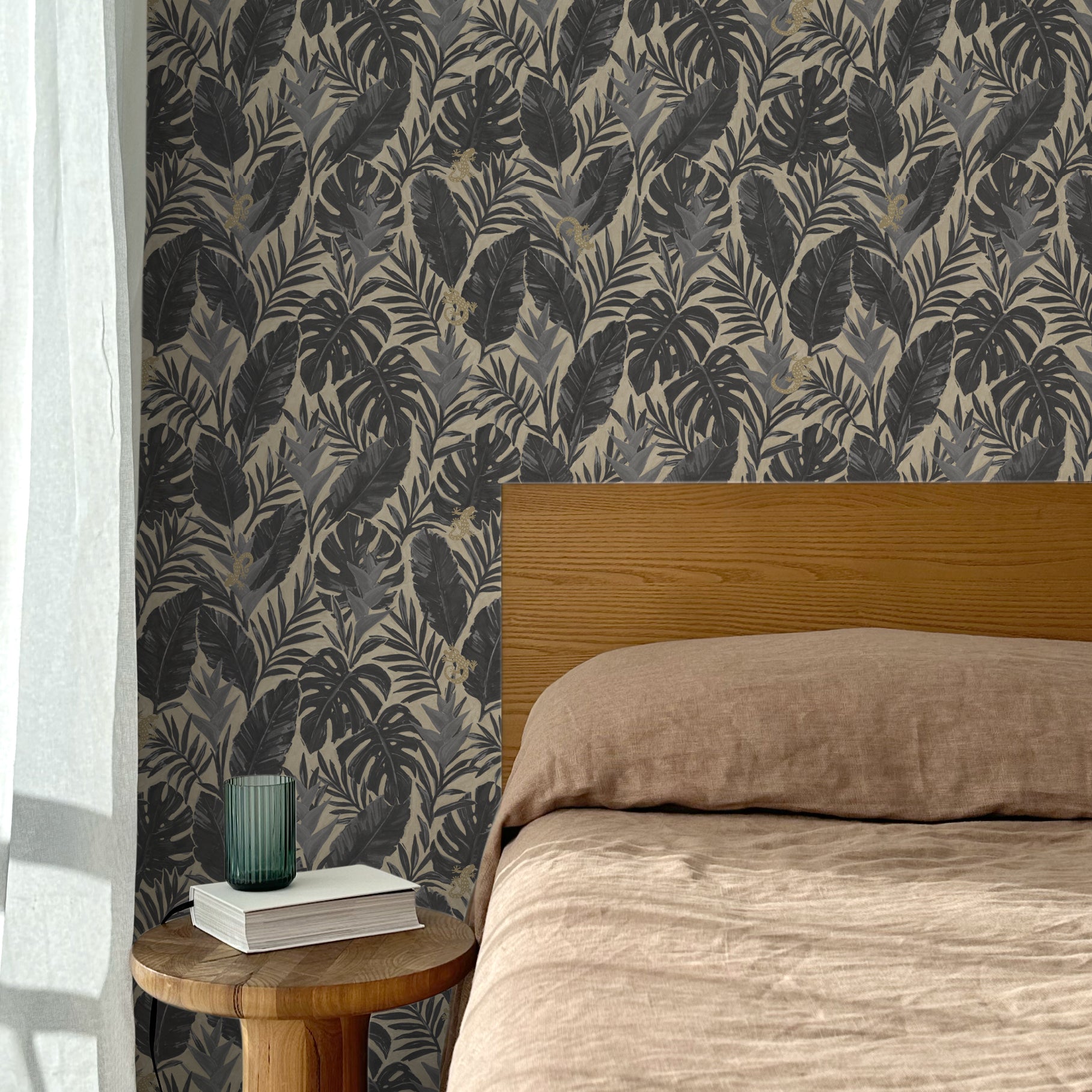 Bed with tan linens and wooden headboard, wooden side table  with white book and green glass, white floor length curtain against a charcoal grey and tan tropical wallpaper. 