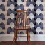 A wooden chair stands in front of a wall papered in a curvy geometric print in shades of white, tan, purple and gray.