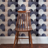 A wooden chair stands in front of a wall papered in a curvy geometric print in shades of white, tan, purple and gray.