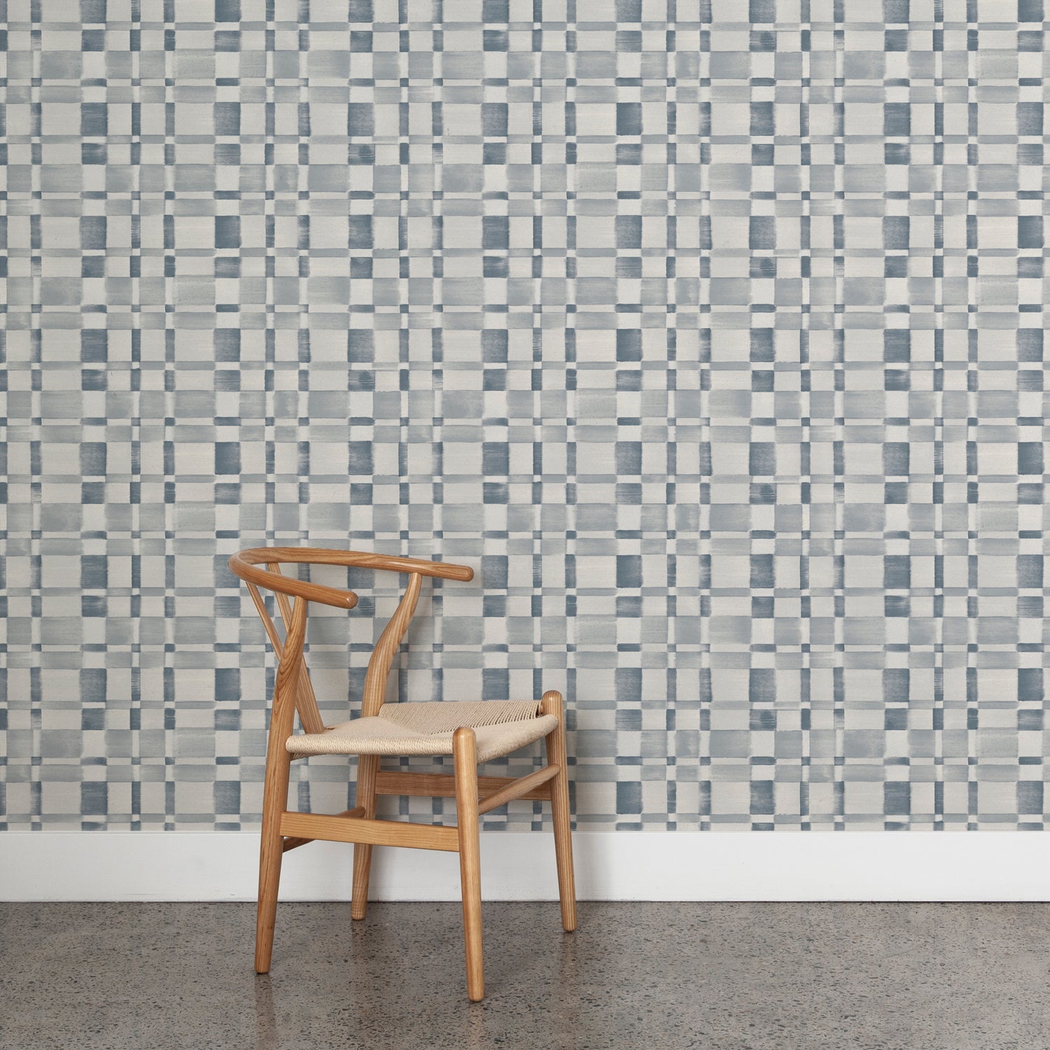 Angled wooden chair with woven seat on concrete floor against wallpaper in a large-scale checked pattern in shades of steel blue and pale grey.