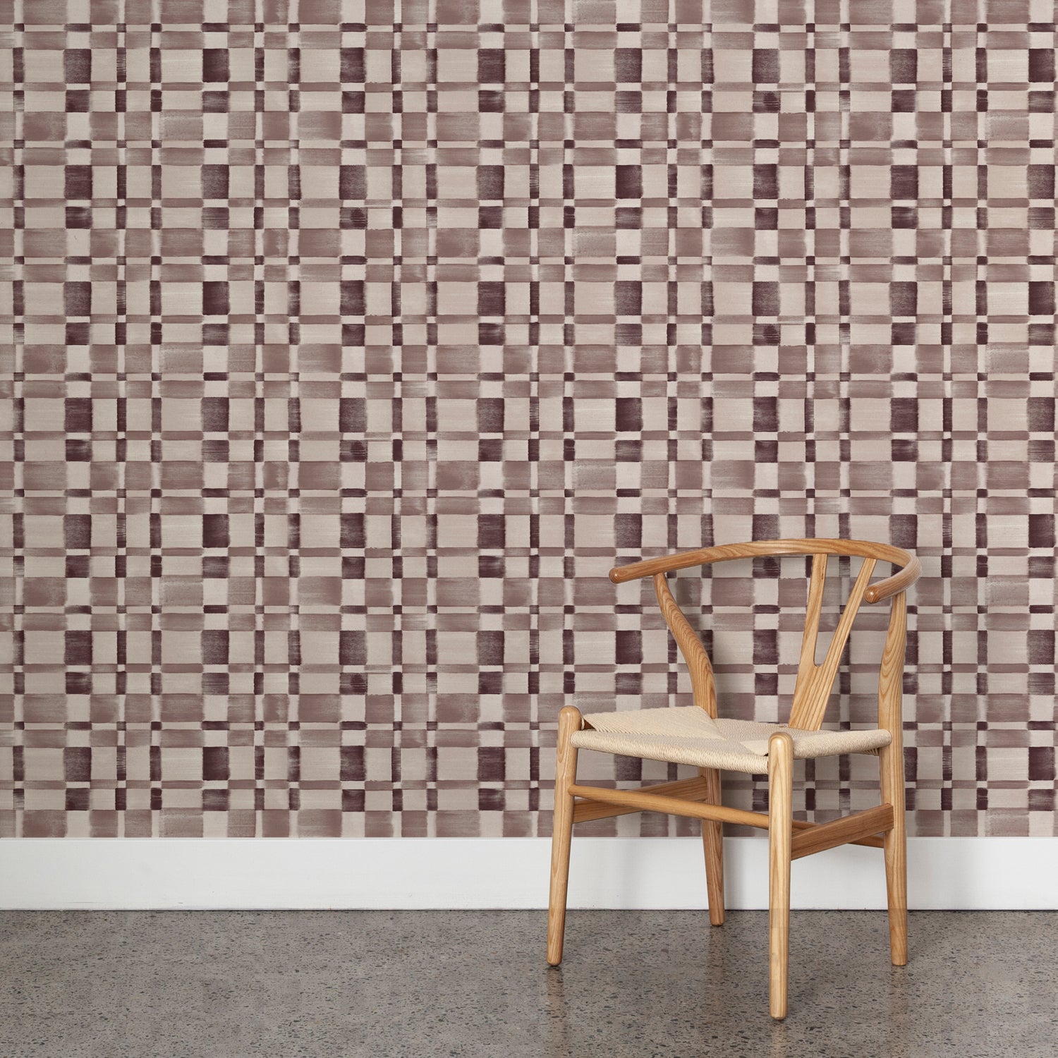 Angled wooden chair with woven seat on concrete floor against wallpaper in a large-scale checked pattern in shades of wine purple and beige.