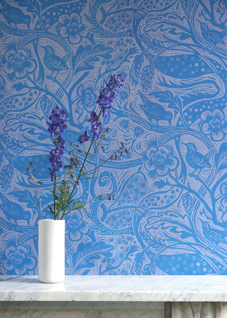 A vase of flowers stands in front of a wall papered in a playful wren and flower print in blue and mauve.