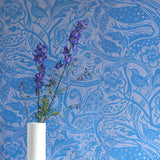 A vase of flowers stands in front of a wall papered in a playful wren and flower print in blue and mauve.