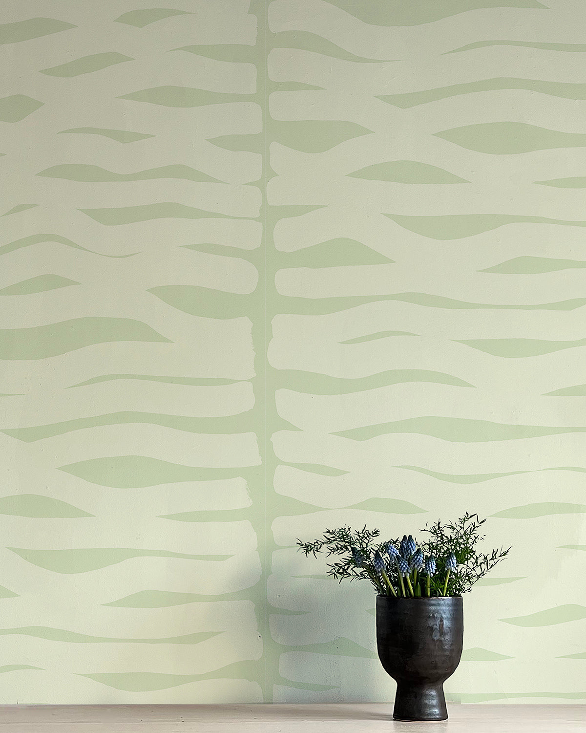 A vase of flowers stands in front of a wall papered in an abstract zigzag pattern in light green on a green field.