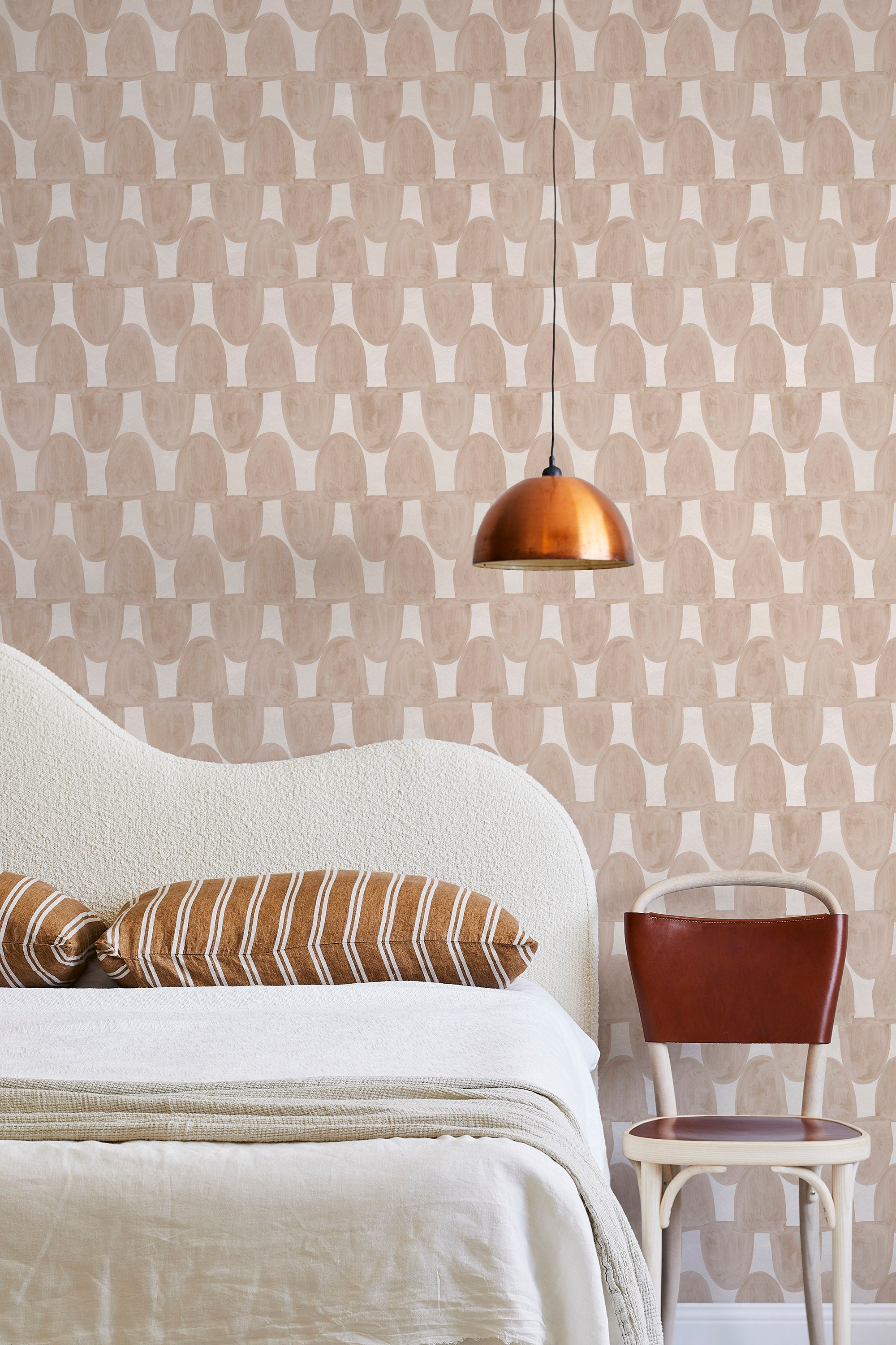 A modernist bed, hanging lamp and chair stand in front of a wall papered in an abstract scalloped print in tan and cream.