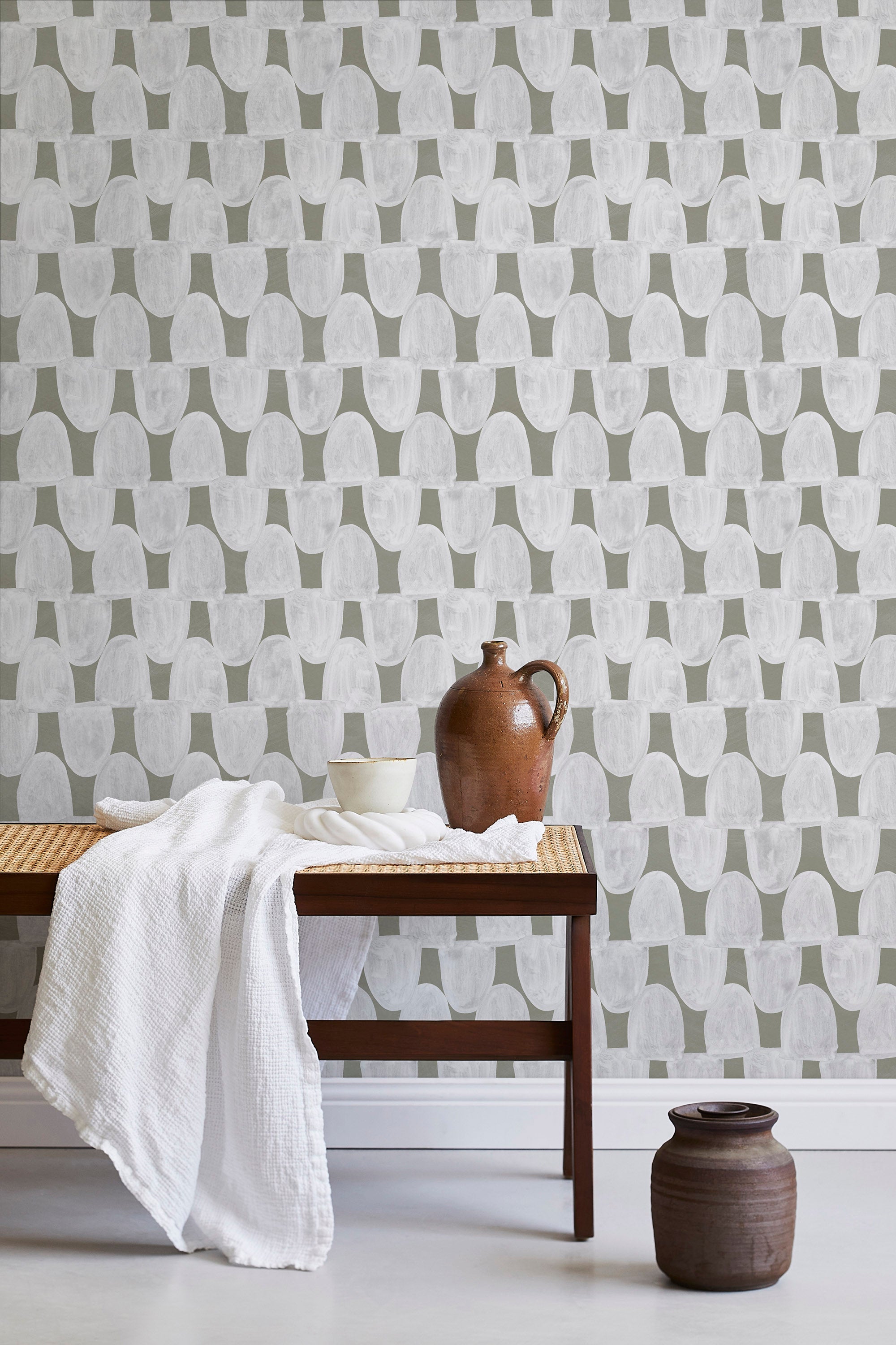 A bench with a towel and clay vases stands in front of a wall papered in an abstract scalloped print in white and olive.