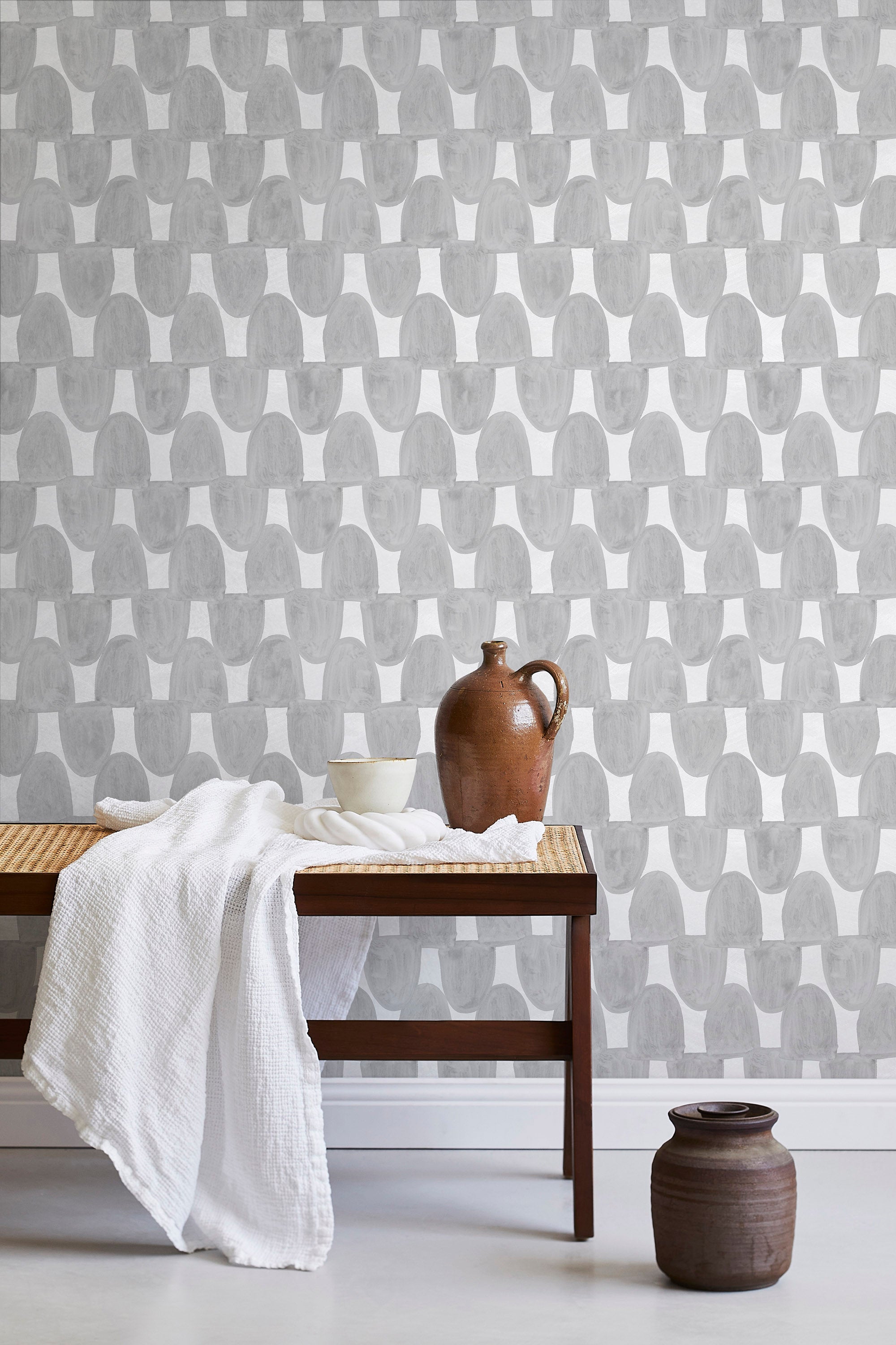 A bench with a towel and clay vases stands in front of a wall papered in an abstract scalloped print in gray and white.