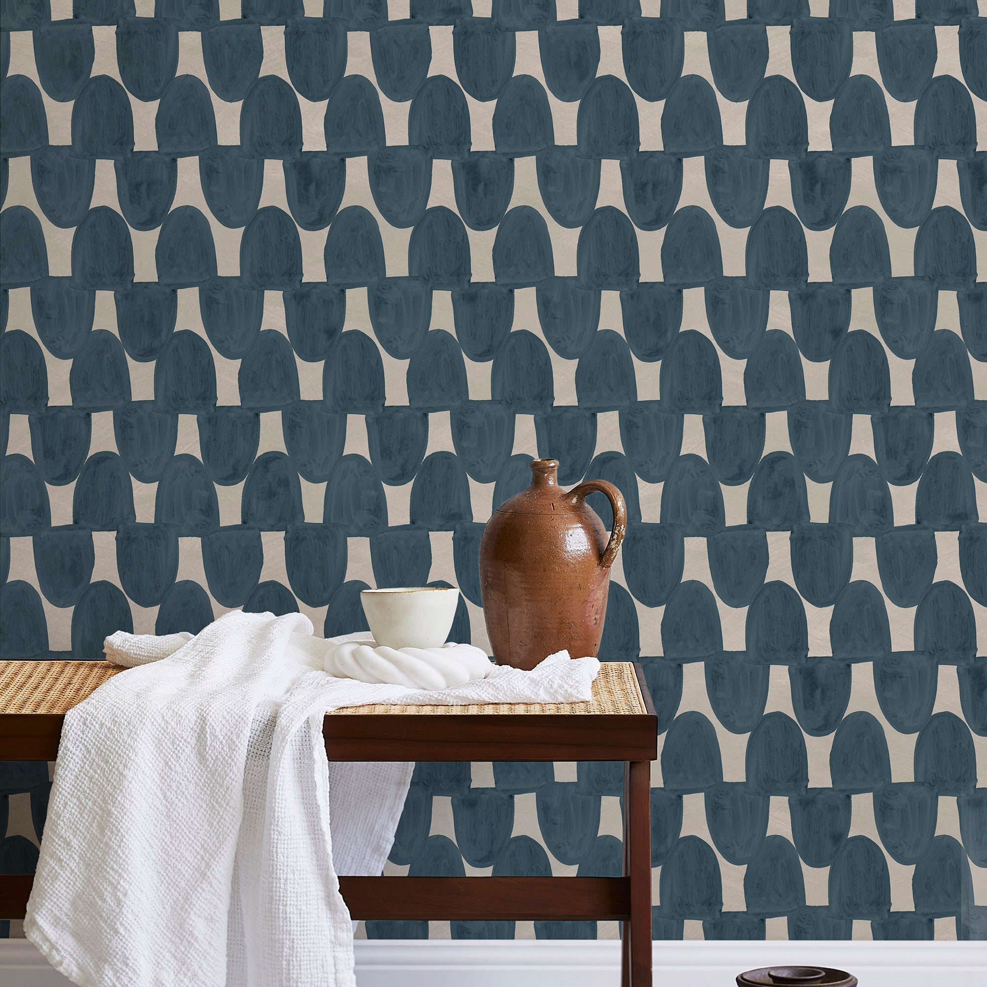 A bench with a towel and clay vases stands in front of a wall papered in an abstract scalloped print in navy and greige.