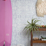 A maximalist room tableau with a purple surfboard and wall papered in a watercolor dyed grid print in purple and turquoise.