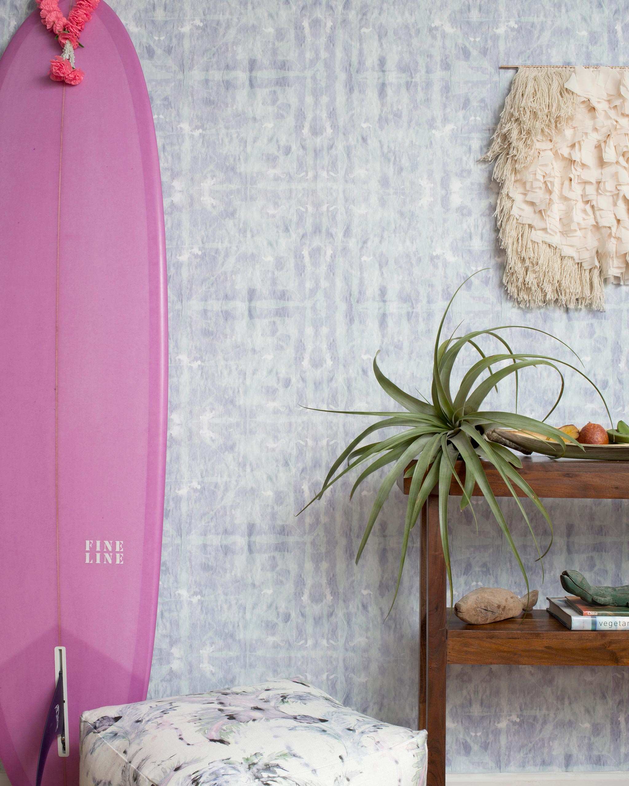 A maximalist room tableau with a purple surfboard and wall papered in a watercolor dyed grid print in purple and turquoise.