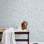 A bench with a towel and clay vases stands in front of a wall papered in a textural checked print in light blue and white.