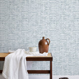 A bench with a towel and clay vases stands in front of a wall papered in a textural checked print in light blue and white.