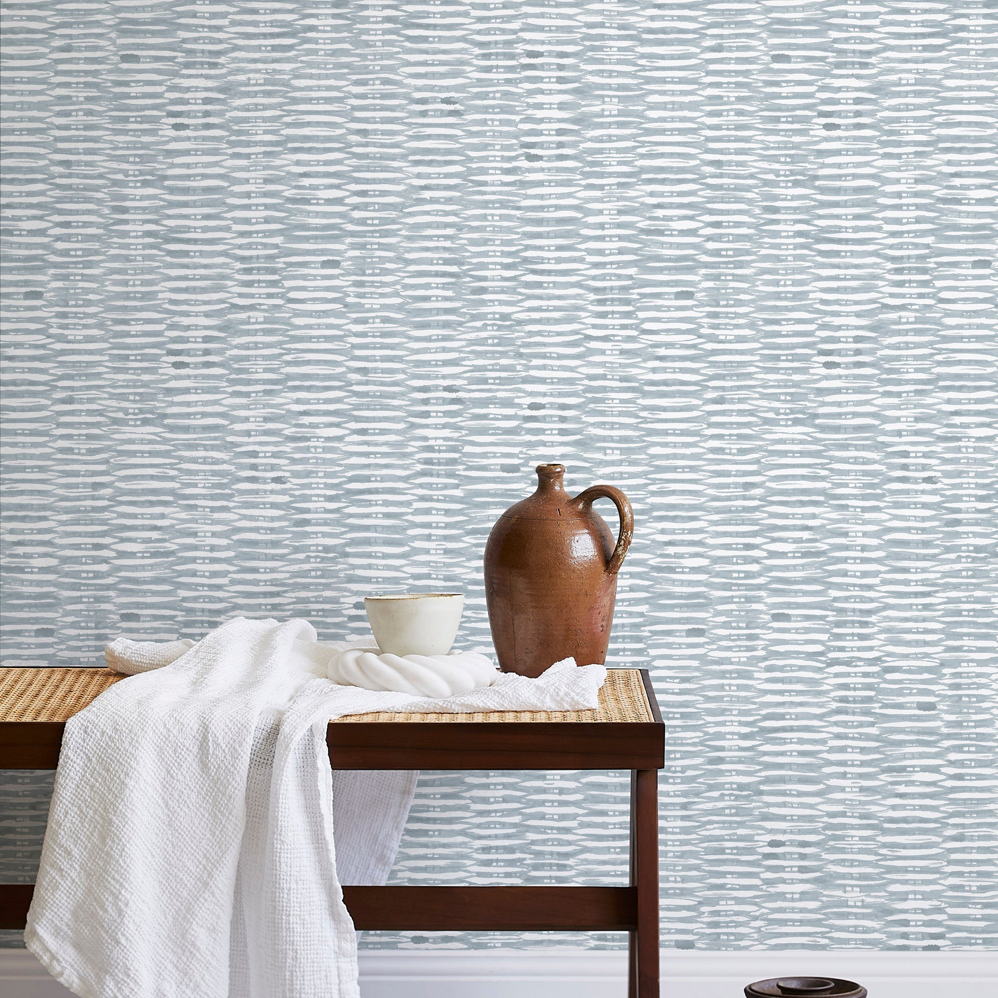 A bench with a towel and clay vases stands in front of a wall papered in a textural checked print in light blue and white.