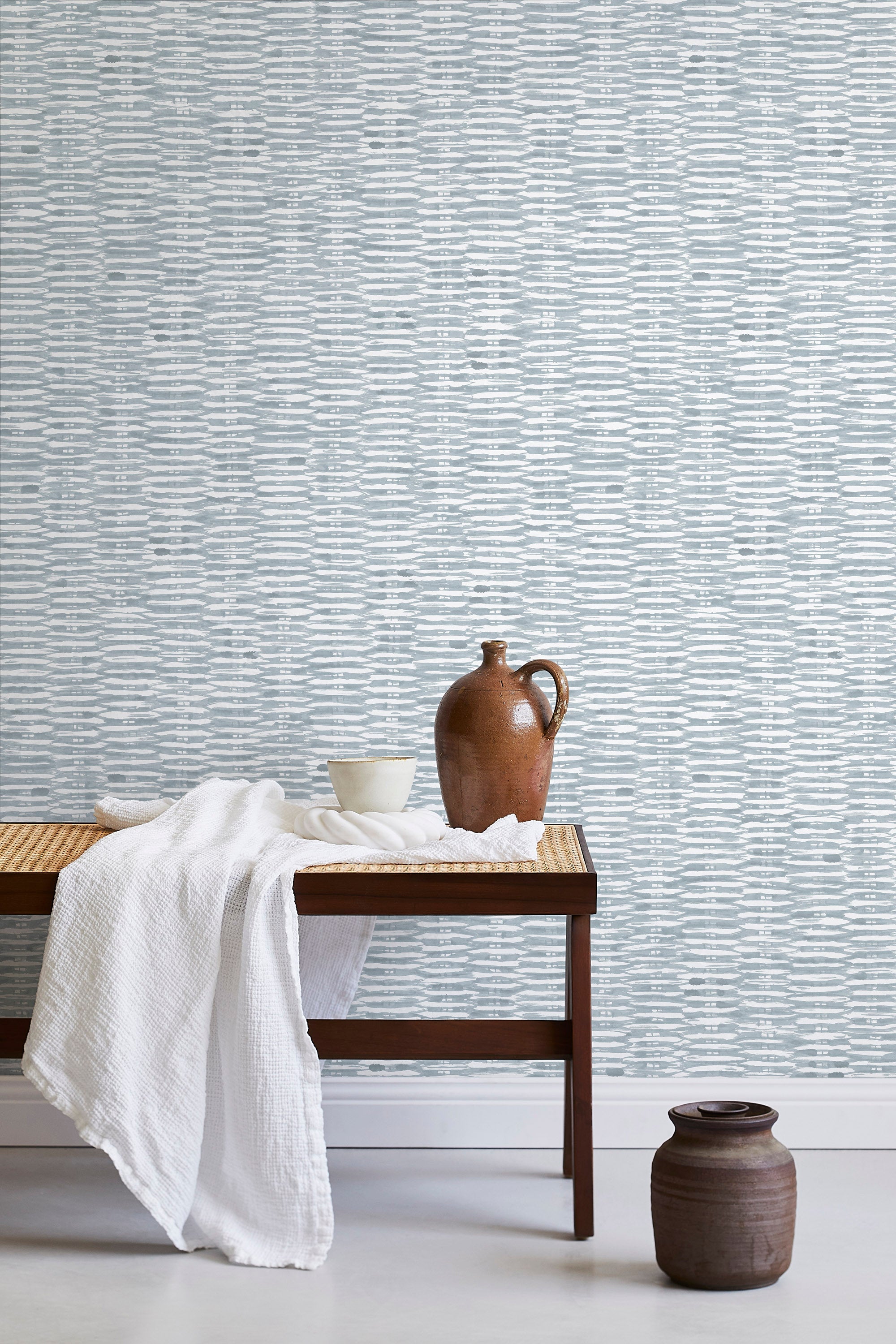A bench with a towel and clay vases stands in front of a wall papered in a textural checked print in light blue and white.