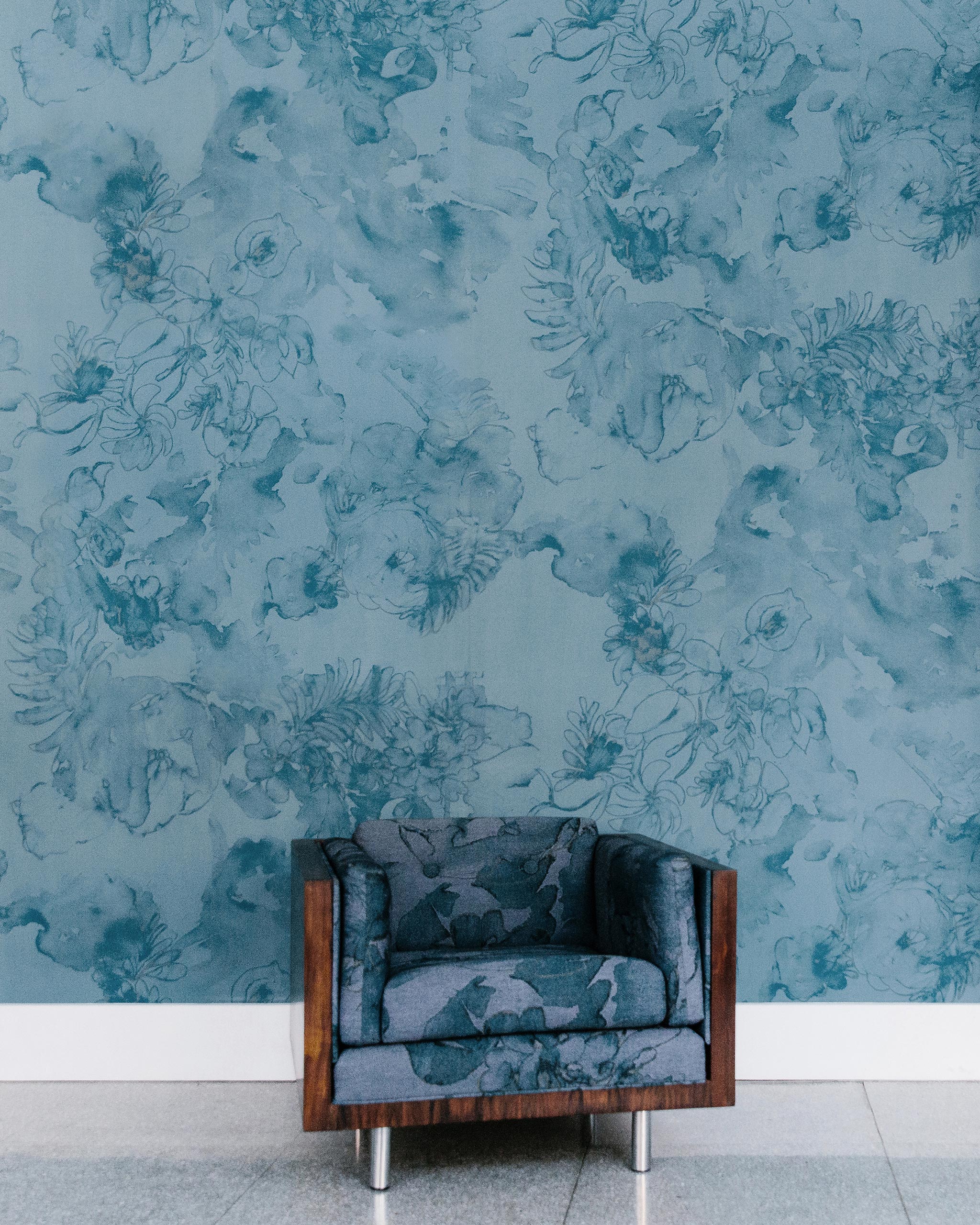 An upholstered armchair stands in front of a wall papered in a painterly floral print in shades of navy and yellow on a blue field.