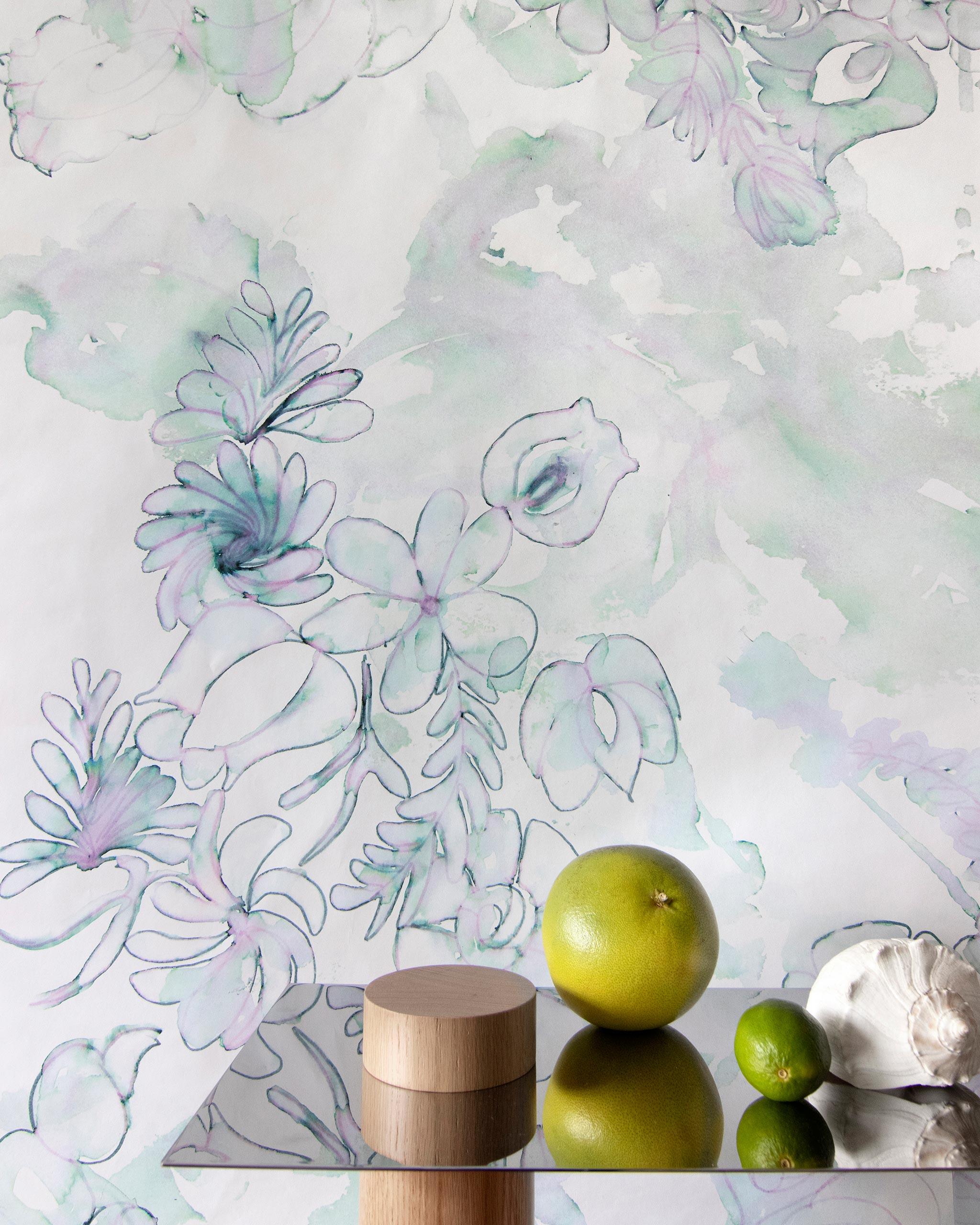A table with fruit and knick knacks stands in front of a wall papered in a painterly floral print in green, purple and white.