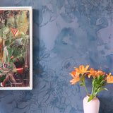 A vase of flowers and painting stand in front of a wall papered in a painterly floral print in navy, tan and blue.