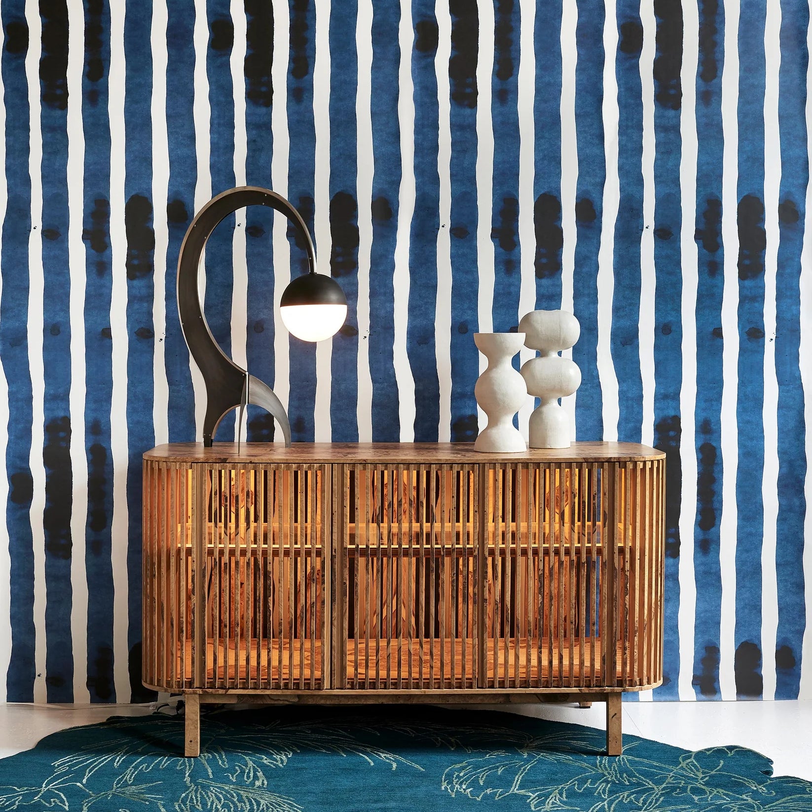 A mid-century modern wooden sideboard cabinet with a slatted bamboo front, styled with a black curved lamp and white ceramic figurines, set against a blue and white striped wallpaper background and a teal rug.