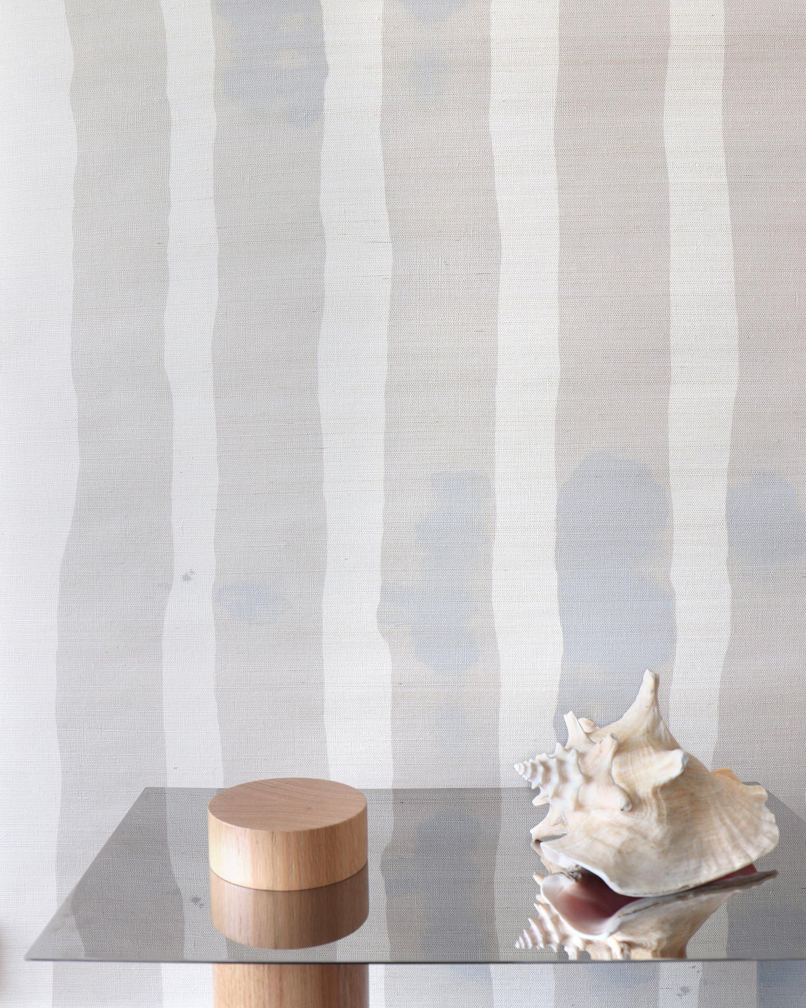 Decorative wood and steel table with conch shell against grey and cream striped grasscloth wallpaper. 