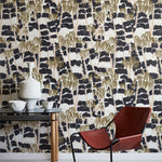 A modernist armchair stands in front of a wall papered in a painterly palm tree print in shades of gold, beige and navy.