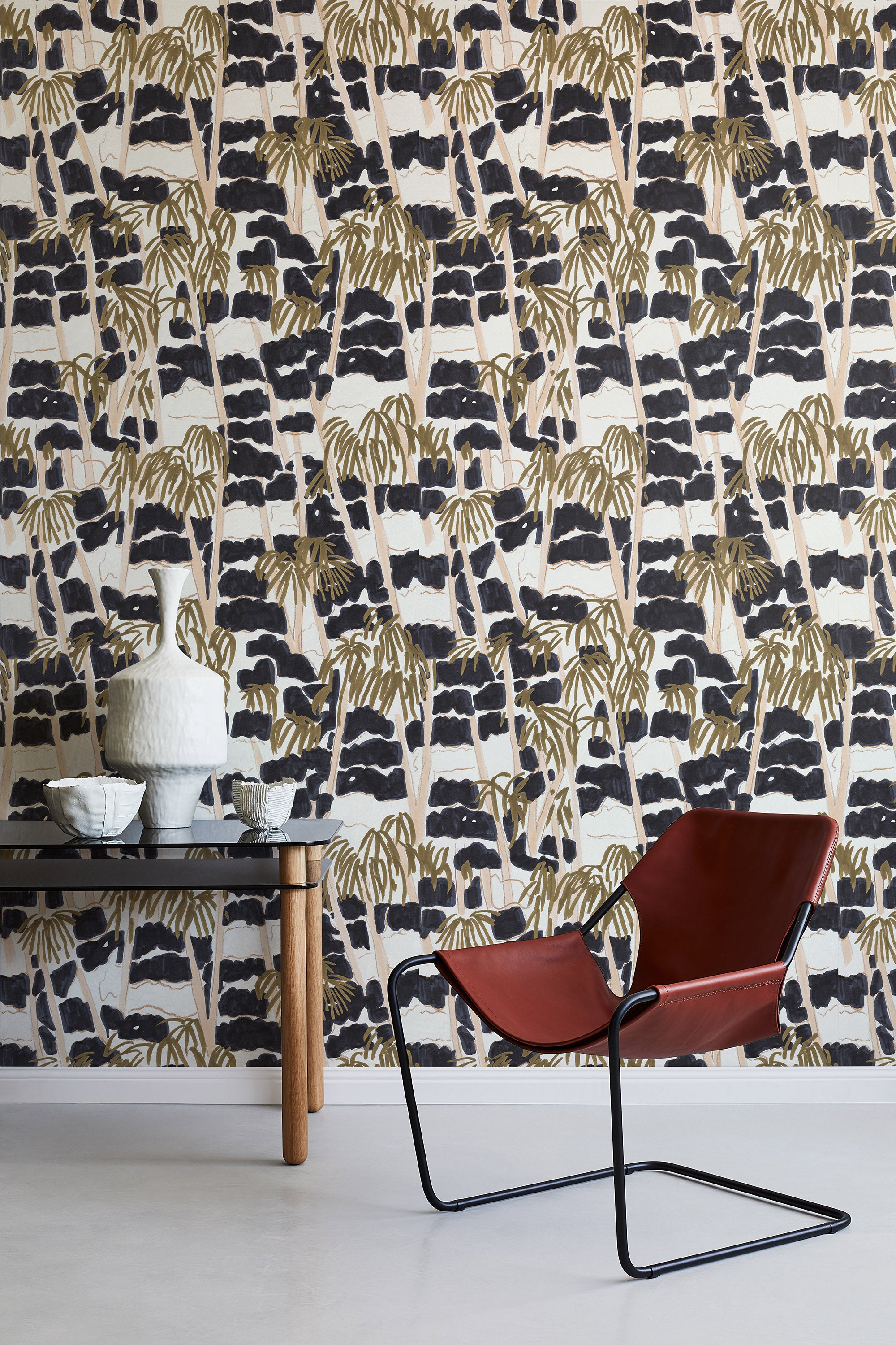 A modernist armchair stands in front of a wall papered in a painterly palm tree print in shades of gold, beige and navy.