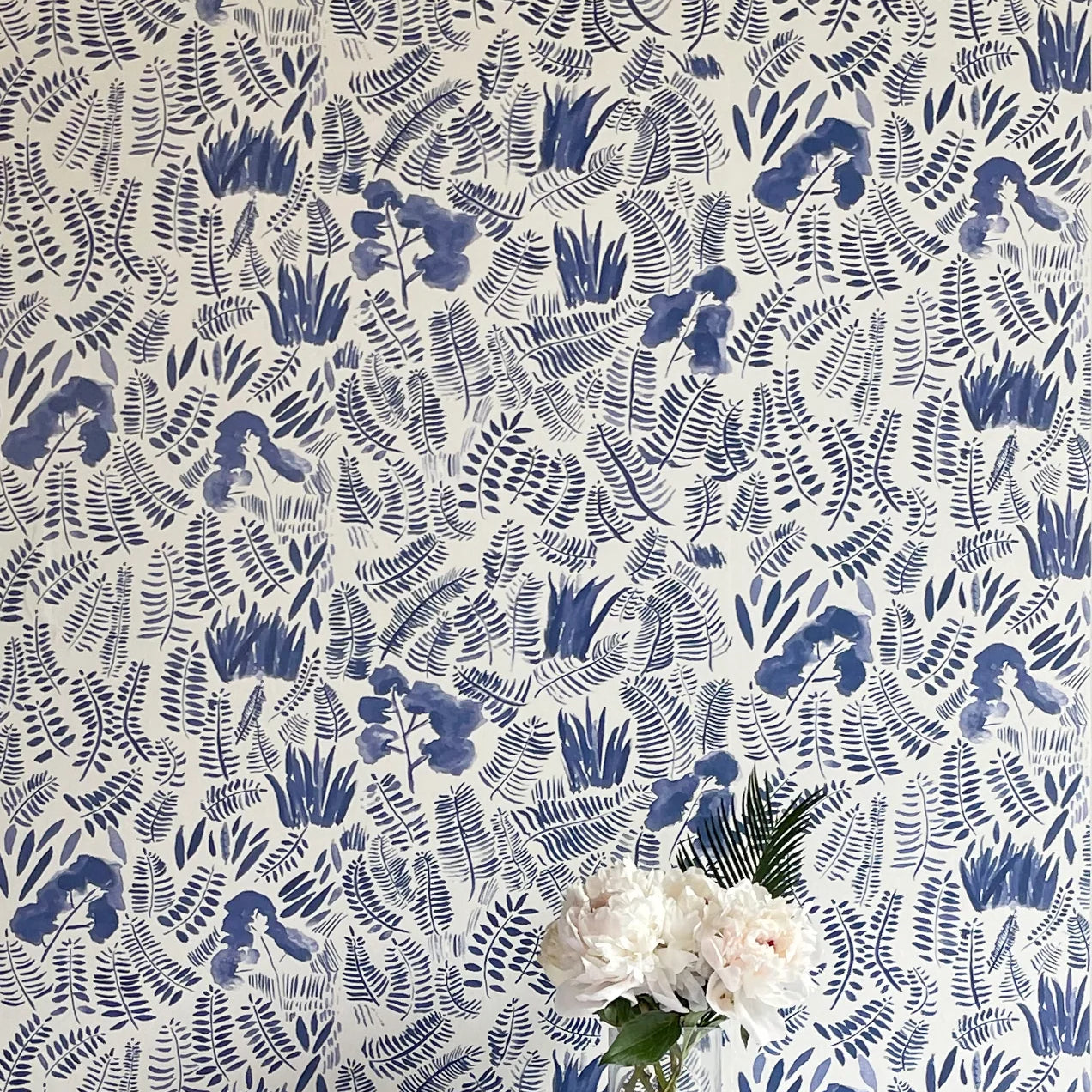 A vase of flowers stands in front of a wall papered in a playful leaf print in shades of blue on a cream field.