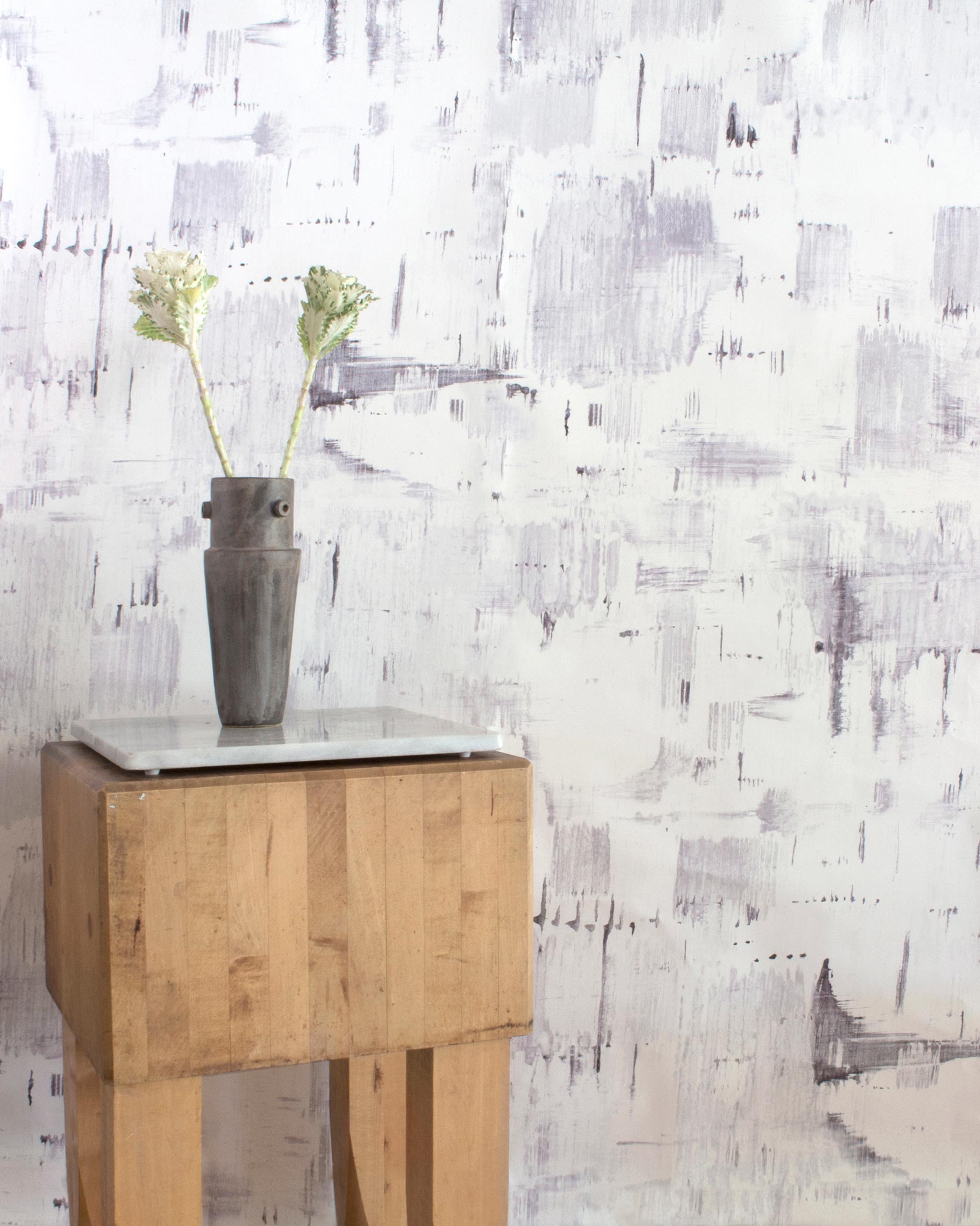 An end table with flowers stands in front of a wall papered in a watercolor textural print in shades of gray on a white field.