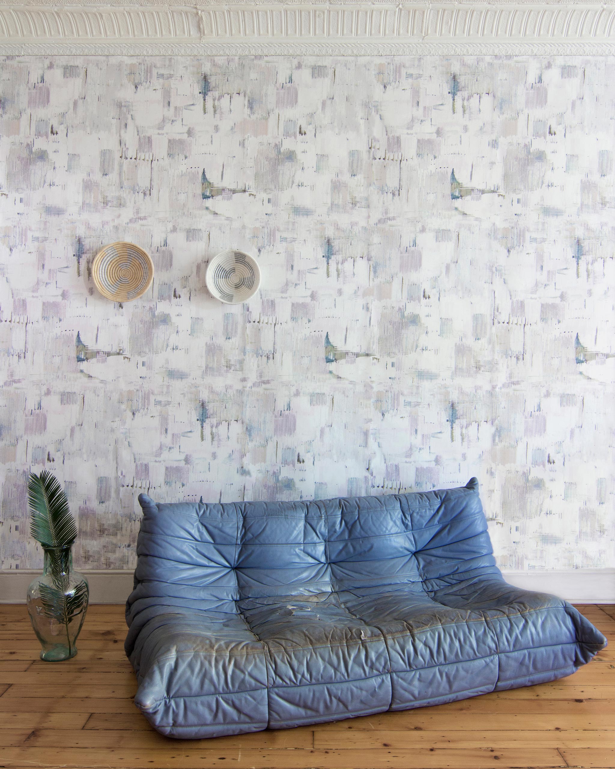 A leather sofa and plant stands in front of a wall papered in a watercolor textural print in green, pink and white.