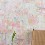 A chair and plant stand in front of a wall papered in a watercolor textural print in shades of pink, green, yellow and white.