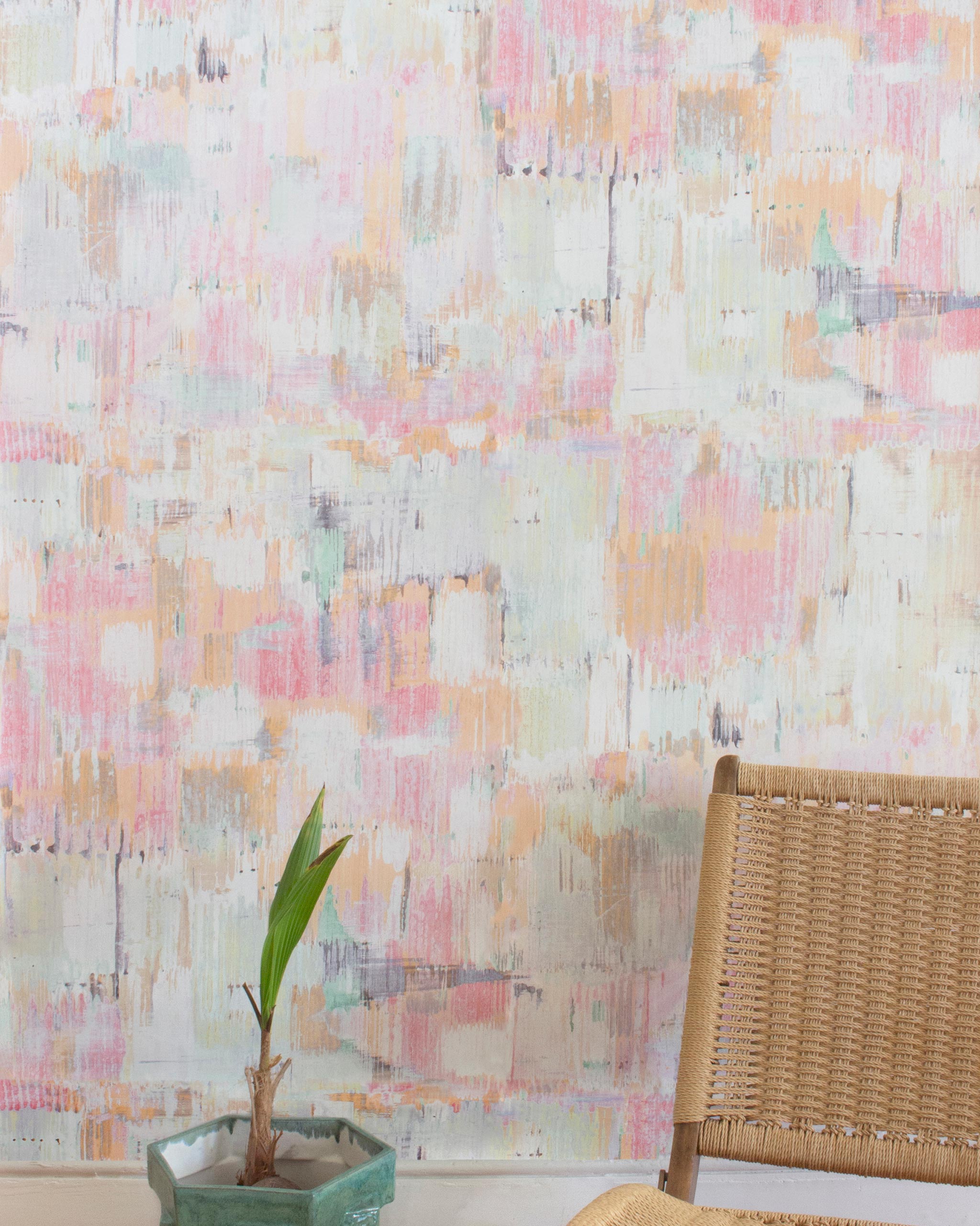 A chair and plant stand in front of a wall papered in a watercolor textural print in shades of pink, green, yellow and white.