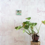 A potted plant on a stand in front of a wall papered in a watercolor textural print in shades of pink, cream and tan.