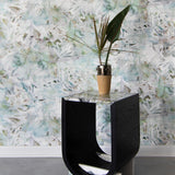 A modernist end table and plant stands in front of a wall papered in a watercolor painted print in green, tan and white.