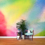 A chair, plant and cat stand in front of a wall covered in an ombré paint splatter print in pink, yellow, blue and green.
