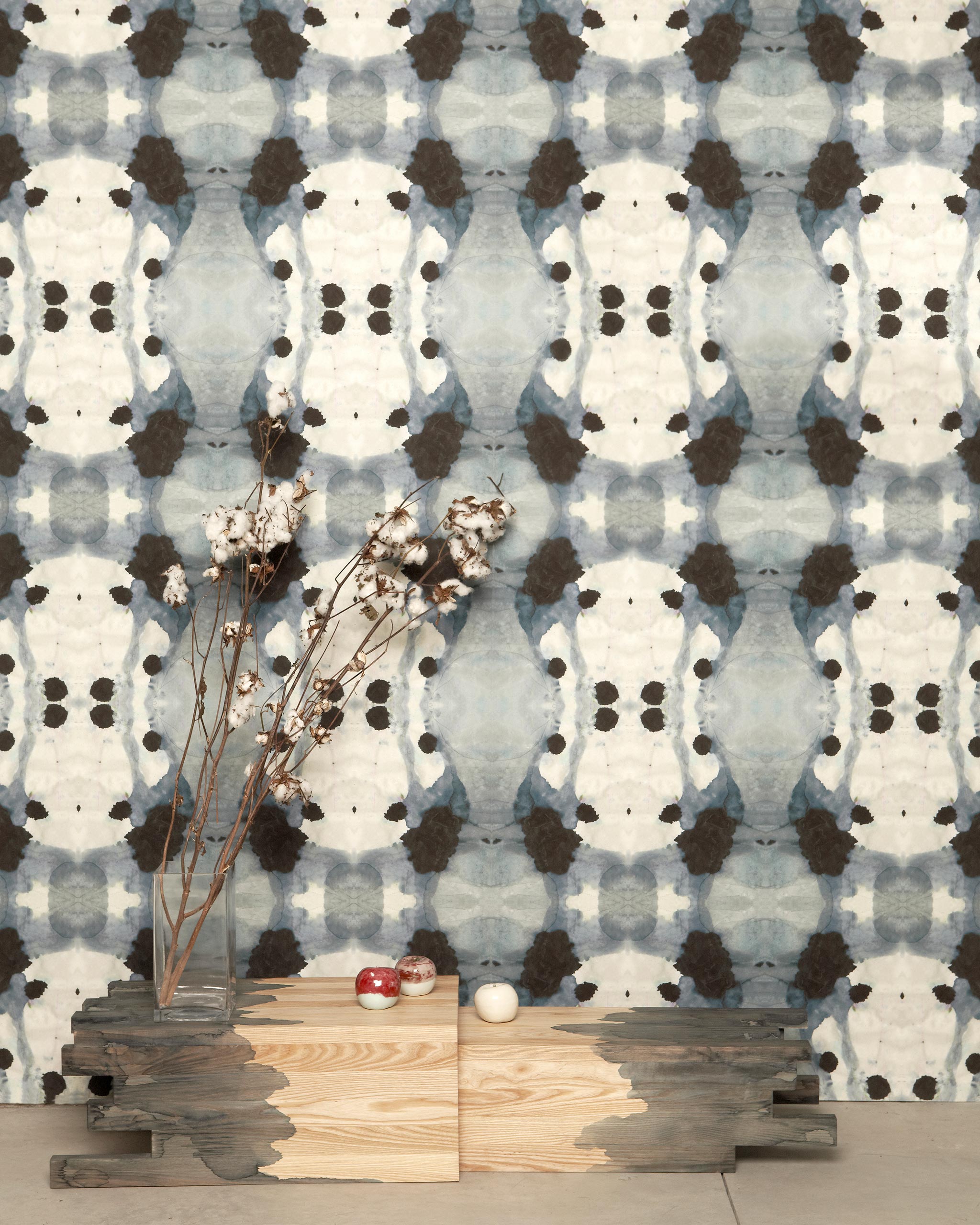 A modernist end table with flowers stands in front of a wall papered in a watercolor ink blot print in charcoal, gray and tan.
