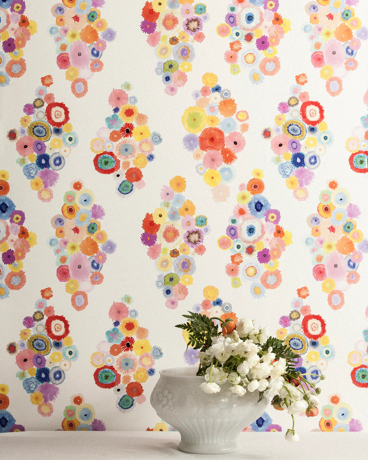 A vase of flowers stands in front of a wall papered in a painterly floral print in multicolor pastels on a white field.