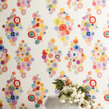 A vase of flowers stands in front of a wall papered in a painterly floral print in multicolor pastels on a white field.