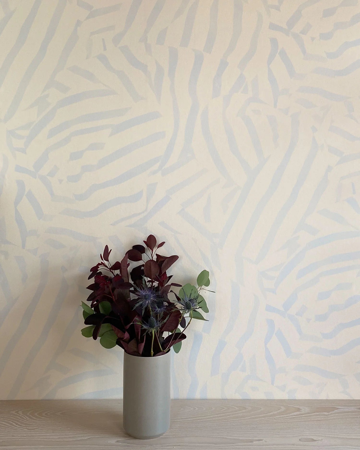 A vase of flowers stands in front of a wall papered in a playful broken stripe print in light blue on a cream field.