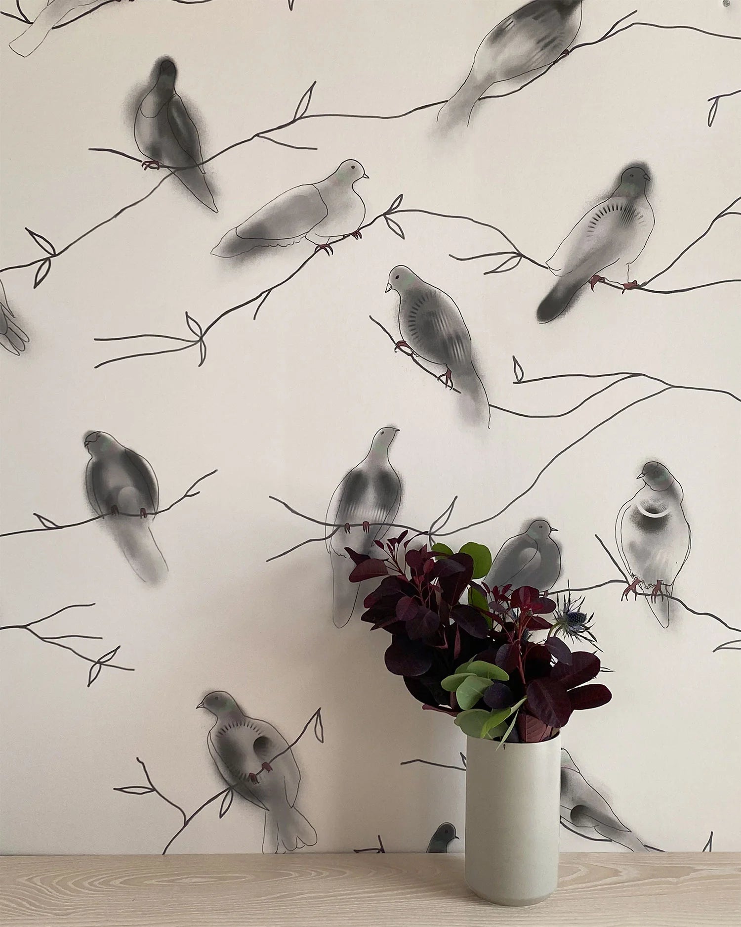 A vase of flowers stands in front of a wall papered in a painterly bird and branch print in black, gray and cream.