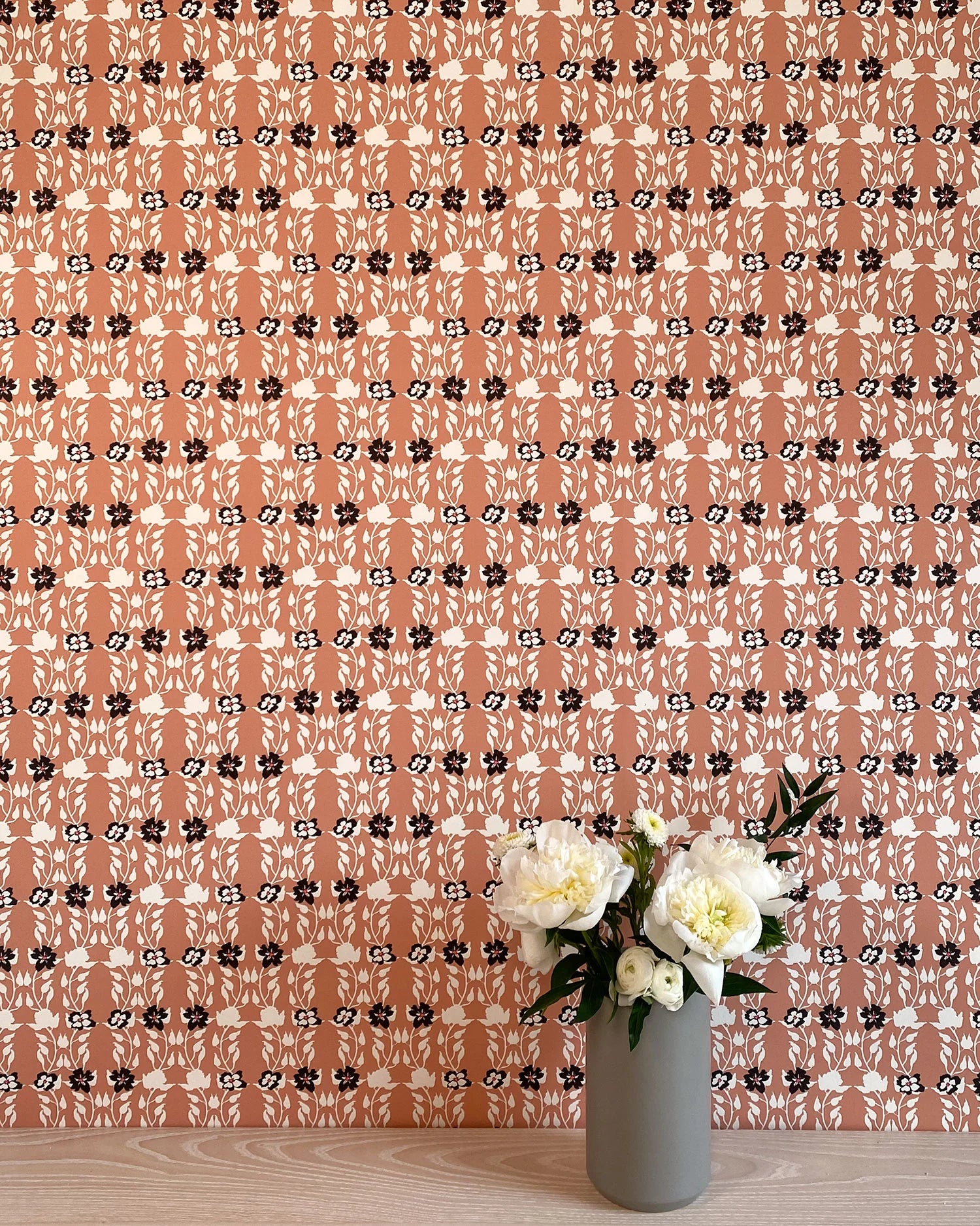 A vase of flowers stands in front of a wall papered in a linear floral print in red, white and black on an orange field.