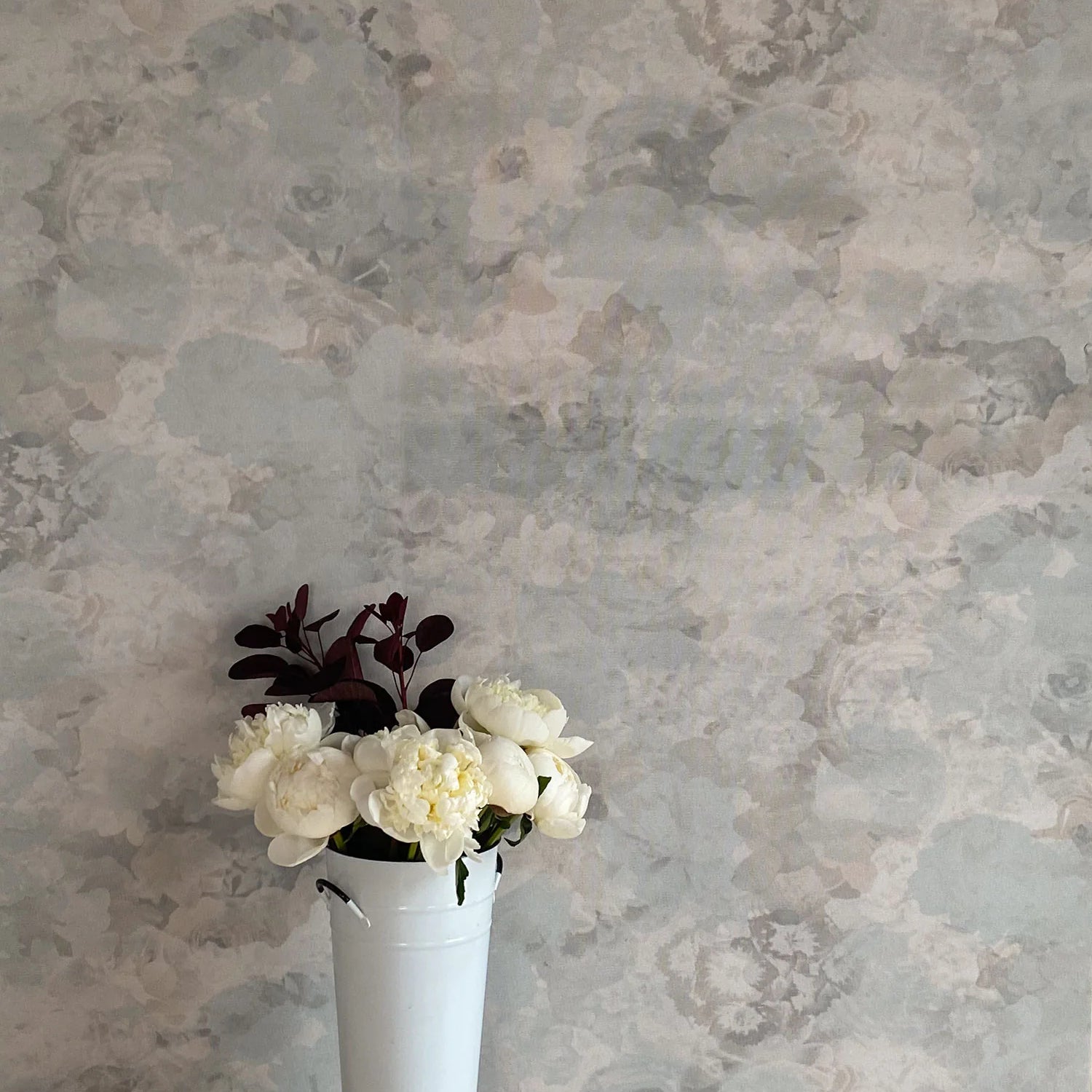 A vase of flowers stands in front of a wall covered in a dense photorealistic floral print in gray.